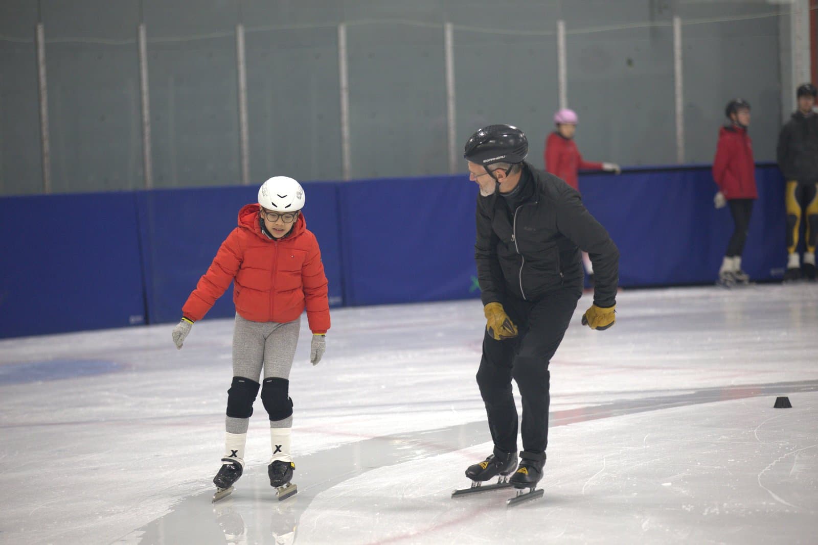 A coach skating alongside a young skater in a red jacket during a Hamilton practice.