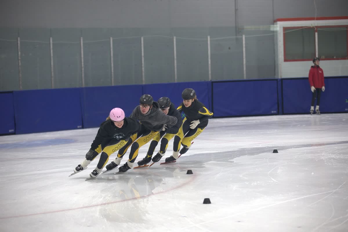 Four Golden Horseshoe skaters in a tight pace line training around a corner with cones on the ice.